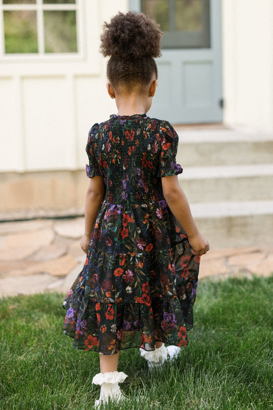A young child with curly hair stands outside, facing away, wearing the Ivy City Co Mini Cordelia Dress in Evergreen Vivid Meadow. The dress features a smocked bodice and puff sleeves. She pairs it with white socks and holds the skirt of her dress while standing on a grassy lawn in front of stone steps and a light-colored house with a blue door.