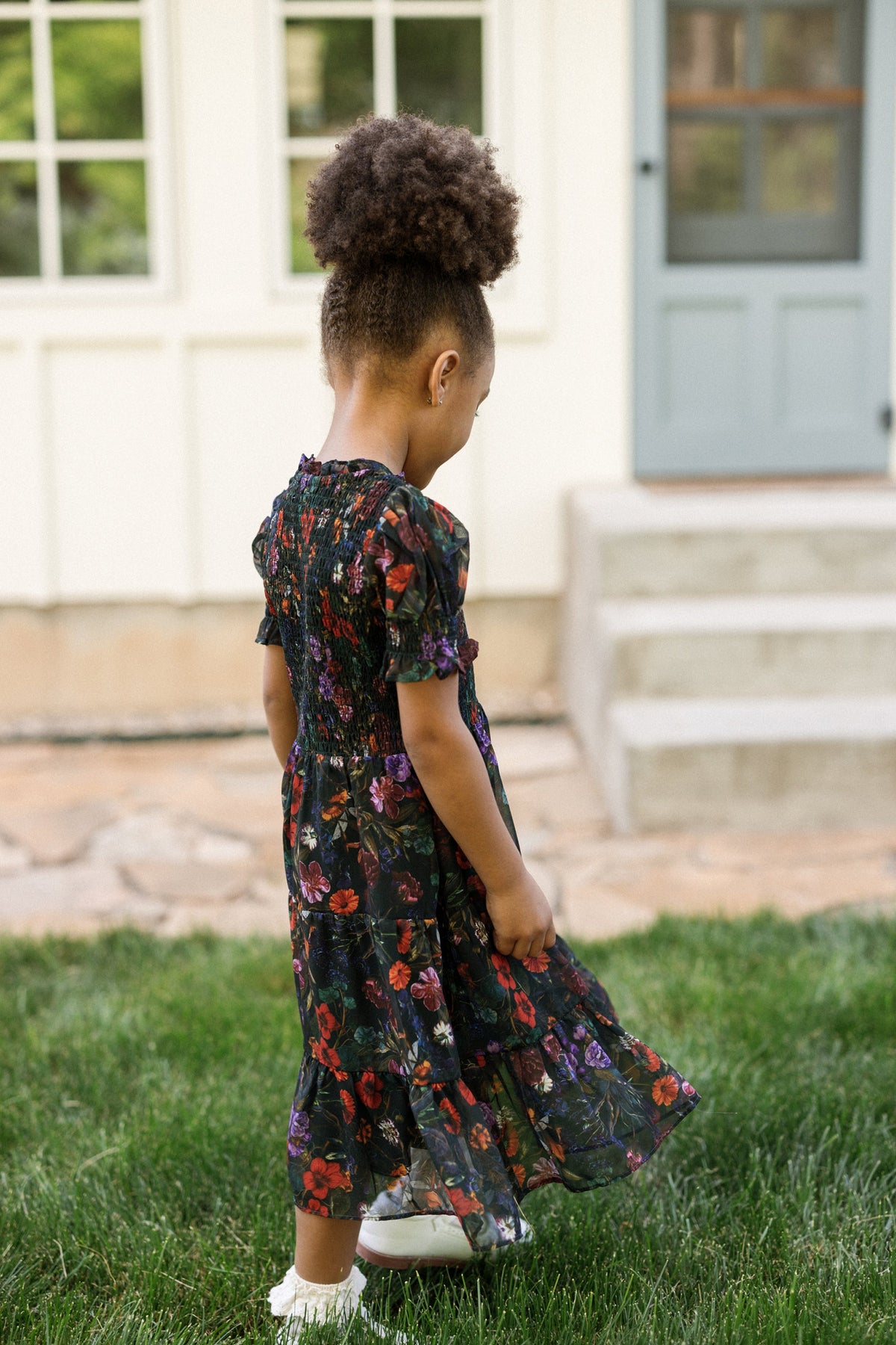 A young girl with curly hair, wearing the Ivy City Co Mini Cordelia Dress in Evergreen Vivid Meadow, featuring a smocked bodice and puff sleeves, stands on the grass looking down, with a house and stone steps in the background.