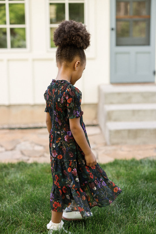 A young girl with curly hair, wearing the Ivy City Co Mini Cordelia Dress in Evergreen Vivid Meadow, featuring a smocked bodice and puff sleeves, stands on the grass looking down, with a house and stone steps in the background.