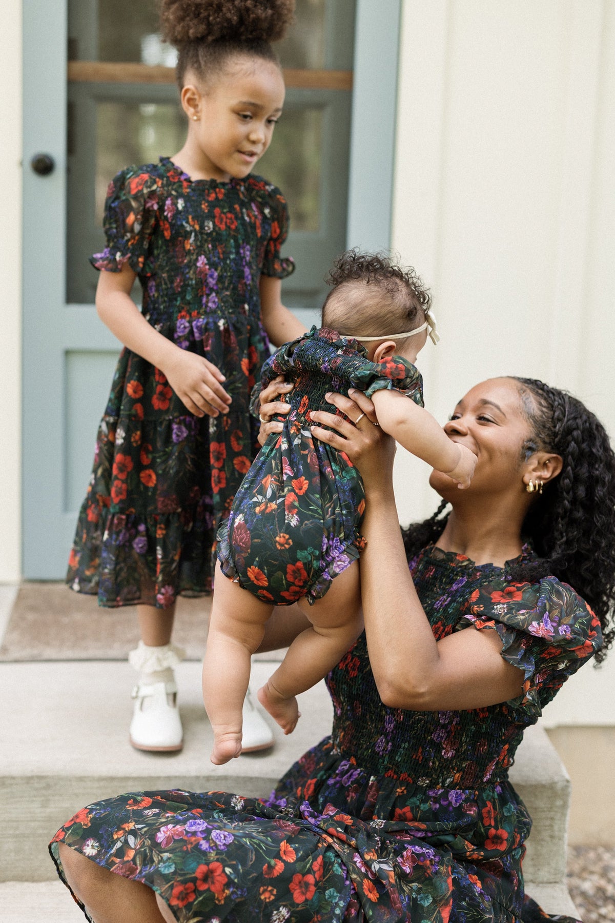 A woman holding a baby and a young girl standing beside them, all wearing matching floral outfits with puff sleeves and smocked bodices, on steps outside a house with a blue door. The young girl is dressed in the Ivy City Co Mini Cordelia Dress in Evergreen Vivid Meadow, perfectly complementing the family’s coordinated look.