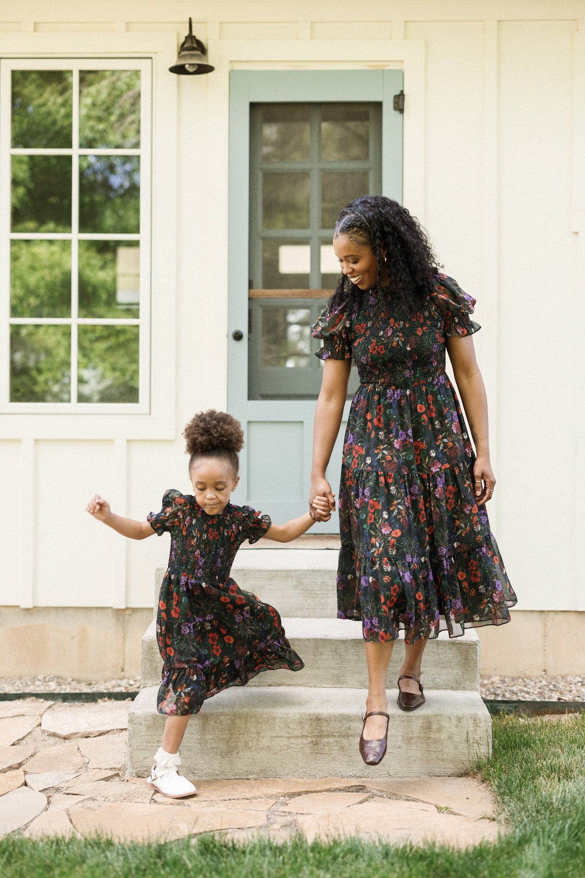 A woman and a young girl, both dressed in matching floral dresses with smocked bodices and puff sleeves from Ivy City Co, hold hands and smile as they step outside a house. The girl gleefully leaps down the front steps, her Mini Cordelia Dress in Evergreen Vivid Meadow fluttering with every joyful bound.