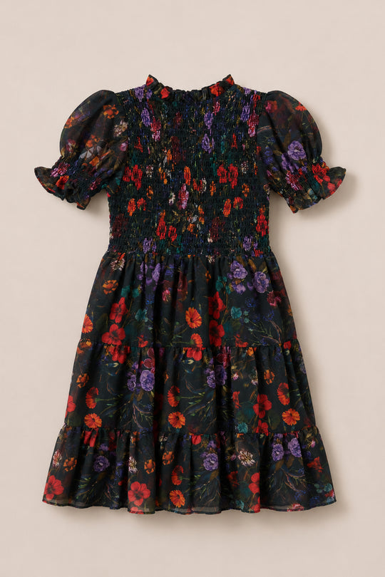 The Ivy City Co Mini Cordelia Dress in Evergreen Vivid Meadow is a short-sleeved, knee-length dress adorned with a vibrant floral print in shades of red, purple, and orange on a dark green background. It features a smocked bodice, puff sleeves, and a ruffled hem.