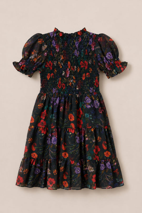 The Ivy City Co Mini Cordelia Dress in Evergreen Vivid Meadow features a smocked bodice and charming puff sleeves, designed in a short-sleeve, knee-length silhouette with a vibrant floral print of red, purple, and green on a classic black background.
