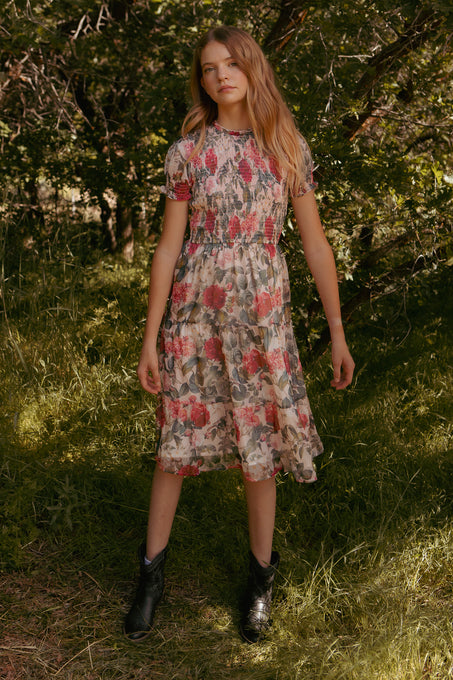 Standing outdoors in a grassy, wooded area, a person wears the Ivy City Co Mini Cordelia Dress in Pink Bloom with black ankle boots. Sunlight filters through the trees, highlighting their long hair.