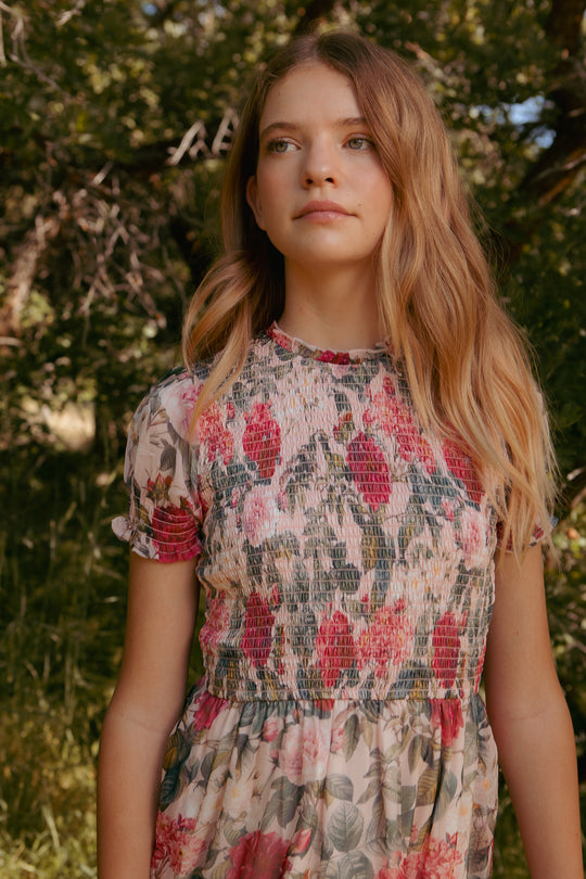 A young woman with long, wavy blond hair stands outdoors in the Mini Cordelia Dress in Pink Bloom by Ivy City Co, looking upward with a serene expression amidst green foliage and tree branches.