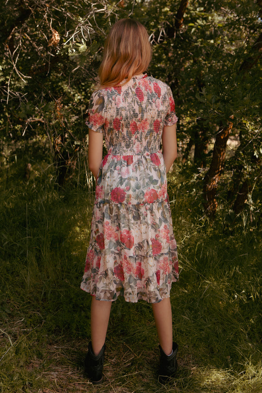 A girl with light brown hair stands outdoors, facing away, wearing the Ivy City Co Mini Cordelia Dress in Pink Bloom and black boots, surrounded by greenery and trees.