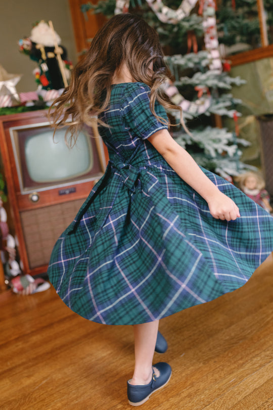 A young girl twirls on a wooden floor in front of a vintage TV and a decorated Christmas tree, wearing the Ivy City Co Mini Cupcake Dress in Emerald Plaid—the perfect look for a cherished family holiday photo.