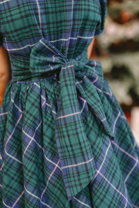 Close-up of the Mini Cupcake Dress in Emerald Plaid by Ivy City Co, highlighting a large, tied bow at the waist. The fabric features a classic green and blue plaid with intersecting white and navy lines, giving this girls’ Christmas dress an elegant look—perfect for capturing memorable family holiday photos. The background is softly blurred.