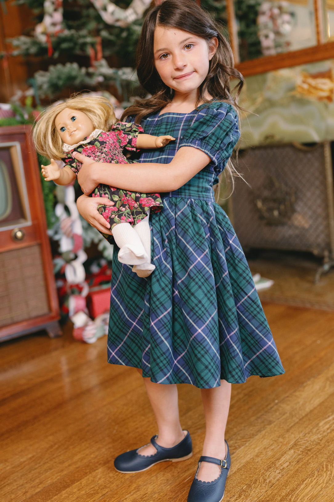 A young girl wearing the Ivy City Co Mini Cupcake Dress in Emerald Plaid holds a blonde-haired doll dressed in a floral outfit. She stands on a wooden floor, surrounded by festive decorations and a Christmas tree in the background.