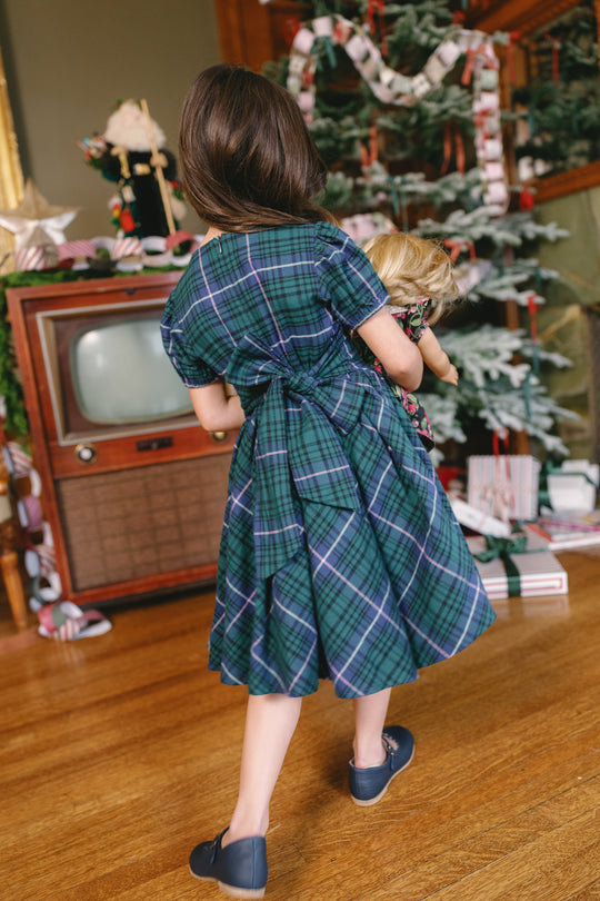 A young girl wearing the Ivy City Co Mini Cupcake Dress in Emerald Plaid holds a doll as she stands in front of a vintage TV beside a decorated Christmas tree adorned with presents and paper chains.