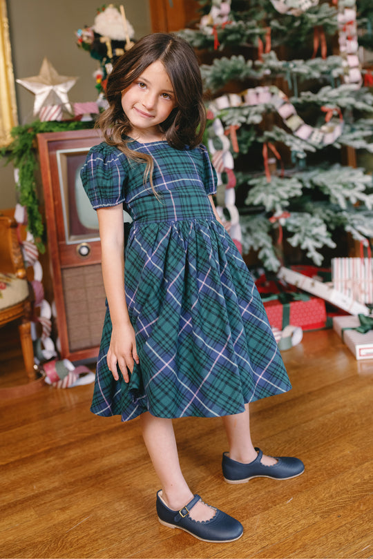 A young girl wearing the Ivy City Co Mini Cupcake Dress in Emerald Plaid poses by a decorated Christmas tree and vintage TV, surrounded by wrapped gifts and festive paper garlands on a wooden floor.