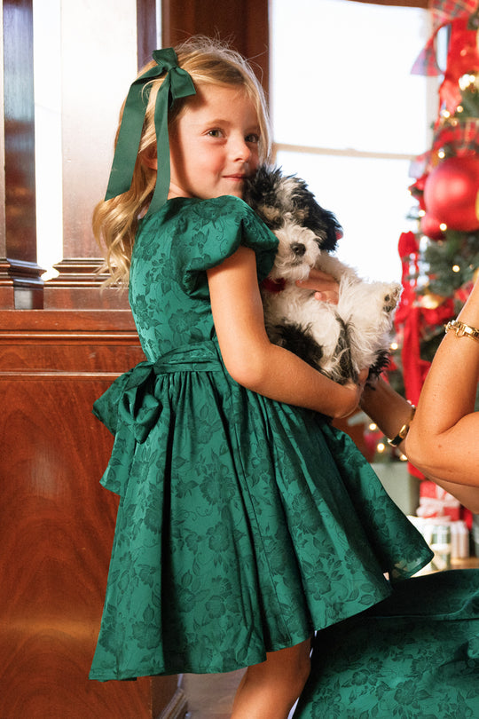 A young girl wears the Ivy City Co Mini Cupcake Dress in Emerald Satin, its bow detail complementing her festive look as she holds a small black and white puppy. She stands indoors by a Christmas tree adorned with red ornaments, perfectly dressed for the holiday party.