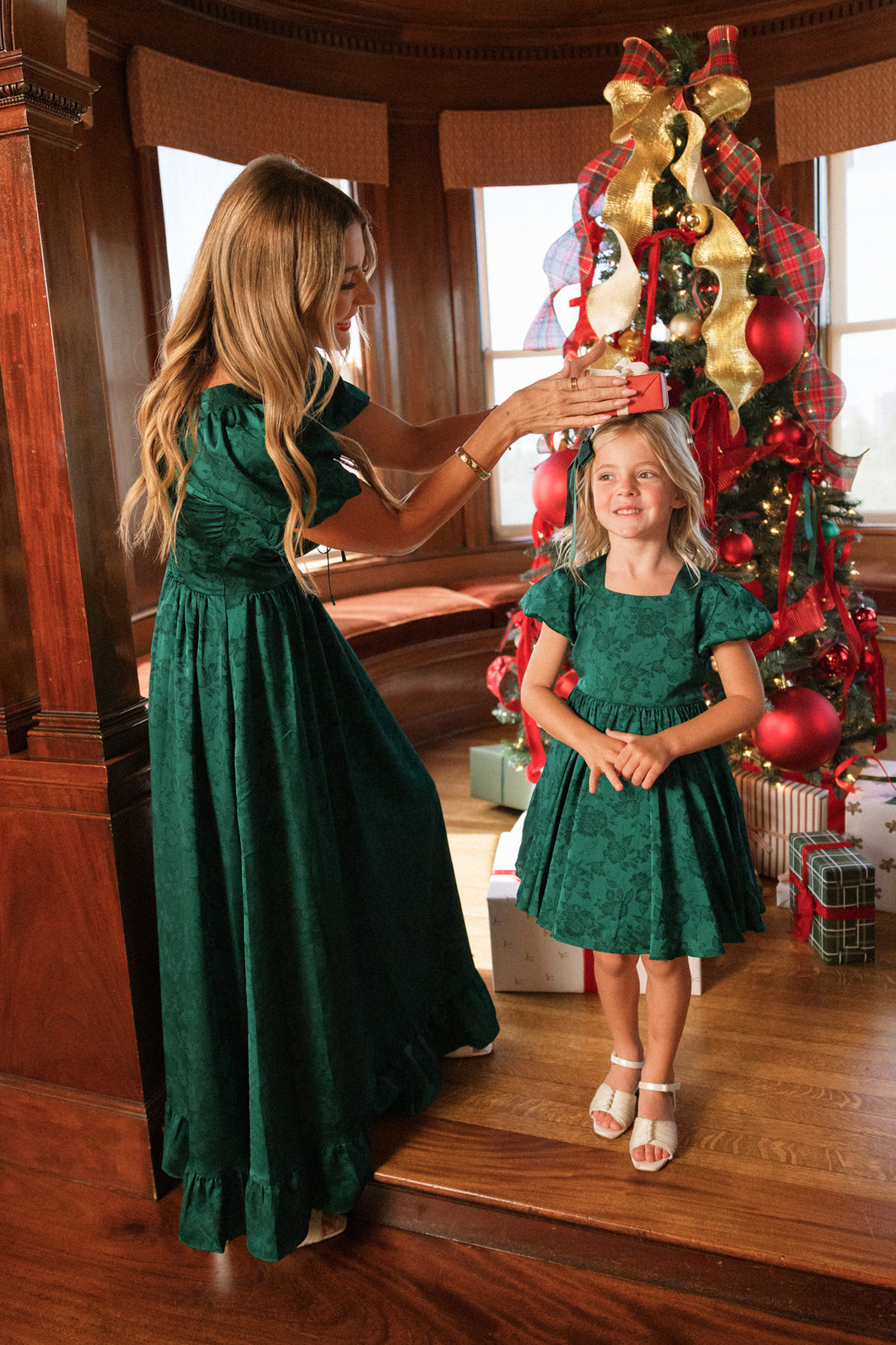 A woman hands a small wrapped gift to a smiling girl in front of a decorated Christmas tree. Both are wearing matching Ivy City Co Mini Cupcake Dress in Emerald Satin, perfect for any holiday party.