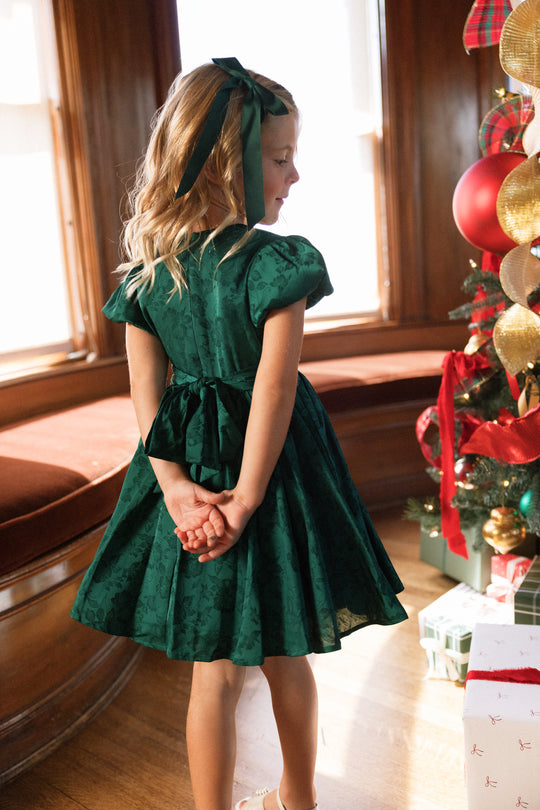 A young girl wearing the Ivy City Co Mini Cupcake Dress in Emerald Satin, complete with a matching bow, stands near a Christmas tree with her hands clasped behind her back, gazing out the window. Presents and festive decorations surround her holiday party dress as warm light fills the room.