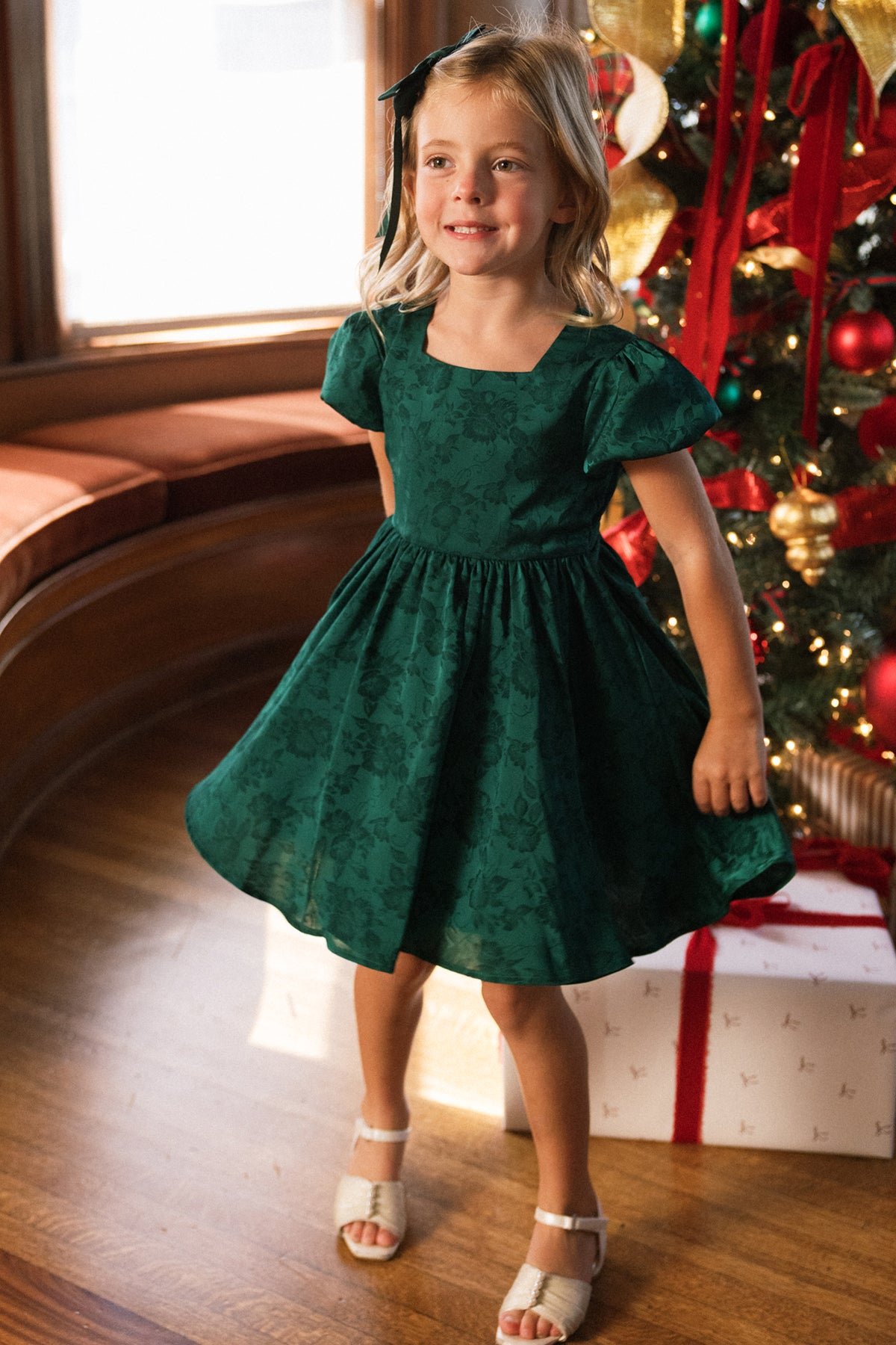 A smiling young girl with blonde hair and a dark green headband is wearing the Ivy City Co Mini Cupcake Dress in Emerald Satin, featuring lovely floral patterns. She stands in a sunlit room beside a decorated Christmas tree and wrapped gifts, perfectly dressed for a festive holiday celebration.