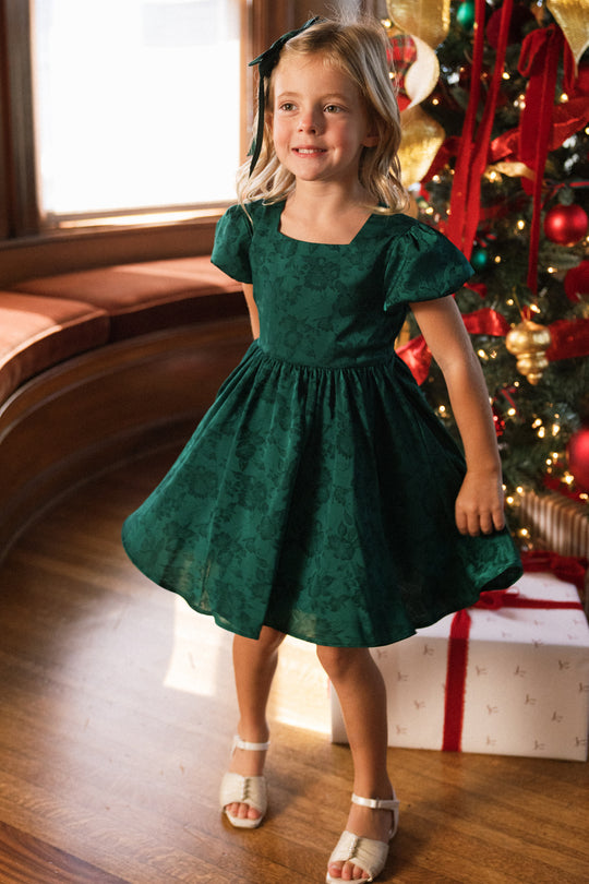 A smiling young girl with blonde hair and a dark green headband is wearing the Ivy City Co Mini Cupcake Dress in Emerald Satin, featuring lovely floral patterns. She stands in a sunlit room beside a decorated Christmas tree and wrapped gifts, perfectly dressed for a festive holiday celebration.