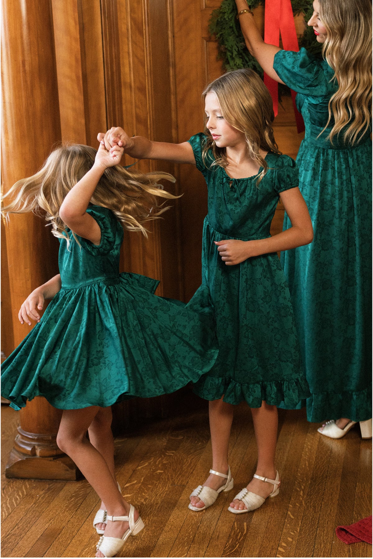Two young girls in matching Ivy City Co Mini Cupcake Dresses in Emerald Satin dance together on a wooden floor, while a woman in a coordinating festive dress hangs a wreath in the background.
