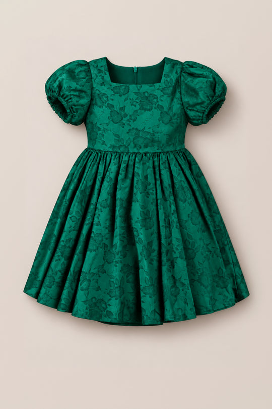Make her holiday shine with the Mini Cupcake Dress in Emerald Satin by Ivy City Co, featuring a fitted bodice, puffed short sleeves, and a full skirt. The luxurious satin fabric is adorned with a subtle floral pattern for festive charm, complete with a back zipper closure. This girls’ festive dress is shown against a plain light beige background.