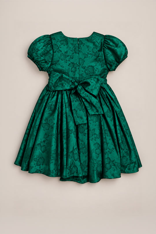 The Mini Cupcake Dress in Emerald Satin by Ivy City Co is a green short-sleeved girls’ festive dress, featuring charming puffed sleeves, a delicate floral pattern, and a large bow at the waist. Its full skirt adds playful elegance, making it perfect as a holiday party dress, and it is beautifully displayed against a plain, light background.