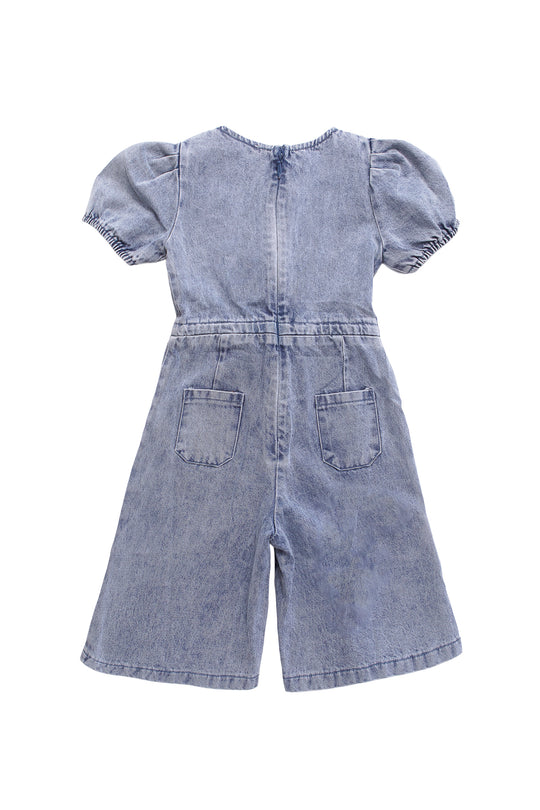 The Ivy City Co Mini Dallas Jumpsuit in Denim is a light blue one-piece for girls with short puff sleeves, wide legs, and two back pockets, displayed flat with the back facing up.