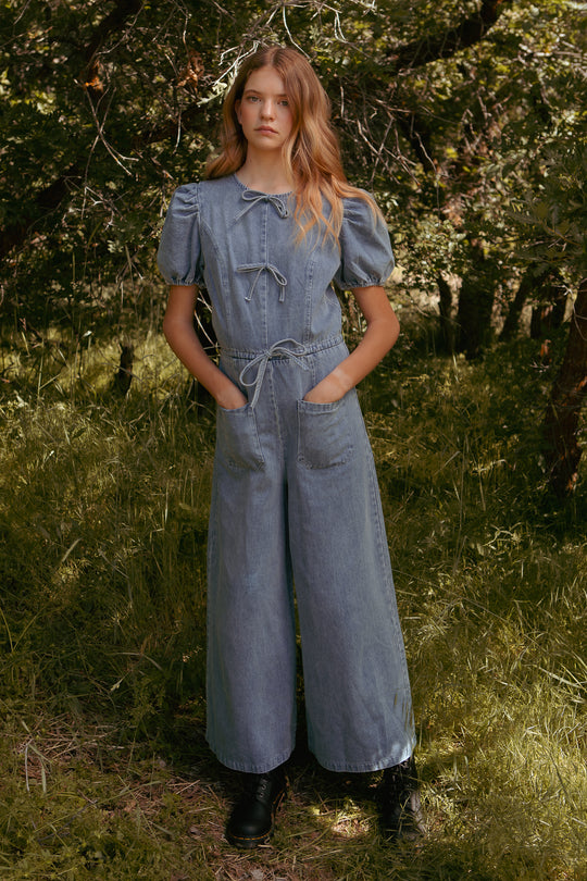 A young woman stands in a grassy, wooded area wearing the Ivy City Co Mini Dallas Jumpsuit in Denim with puff sleeves and wide legs. She has long, wavy hair, black boots, and her hands in the jumpsuit’s front pockets.