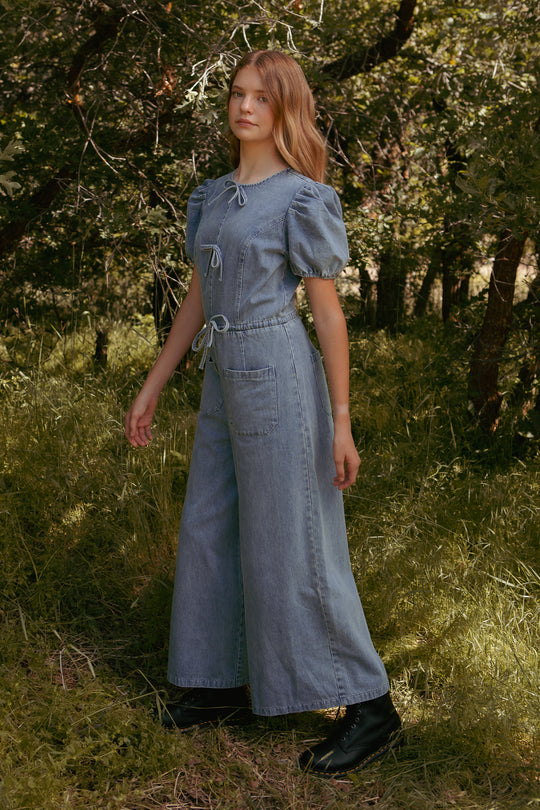 A young woman stands on grass among trees, wearing the Ivy City Co Mini Dallas Jumpsuit in Denim with puffed sleeves and black boots. She gazes to the side with a relaxed look, effortlessly showcasing this trendy one-piece style.