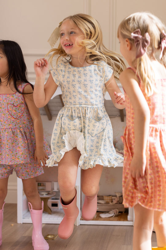 Three young girls wearing Ivy City Co Mini Dream Play Dress in Ivory Porcelain Fleur and rain boots joyfully jump indoors on a light wood floor, with toys and a play kitchen visible in the background.