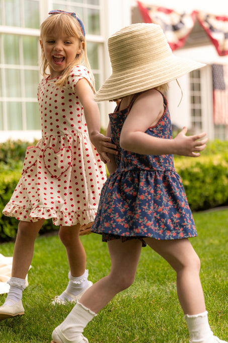 Two young girls dance on the lawn. One twirls in Ivy City Co’s Mini Dream Play Dress in Red Polka Dot, while the other wears a blue floral summer dress with a straw hat. A house behind them is decorated with patriotic bunting.