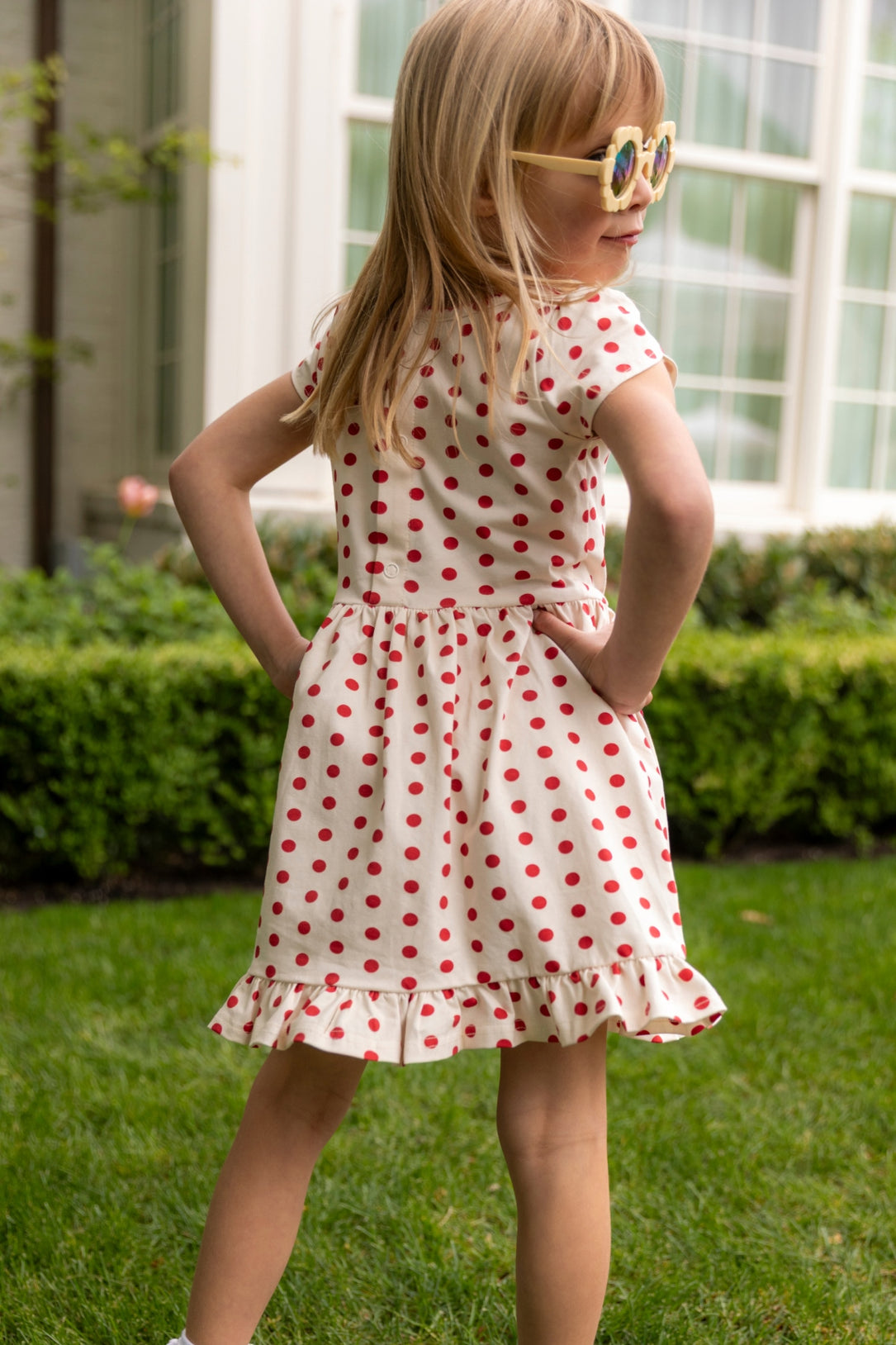 A young girl in the Mini Dream Play Dress in Red Polka Dot by Ivy City Co stands on grass, hands on hips, wearing sunglasses and facing away from the camera before a building with large windows.