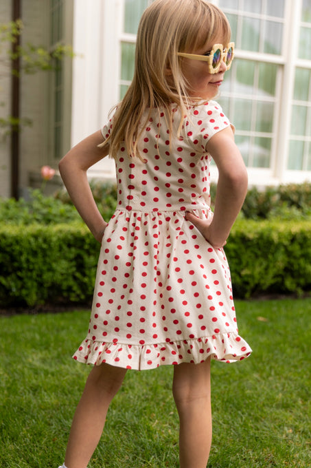 A young girl in the Mini Dream Play Dress in Red Polka Dot by Ivy City Co stands on grass, hands on hips, wearing sunglasses and facing away from the camera before a building with large windows.