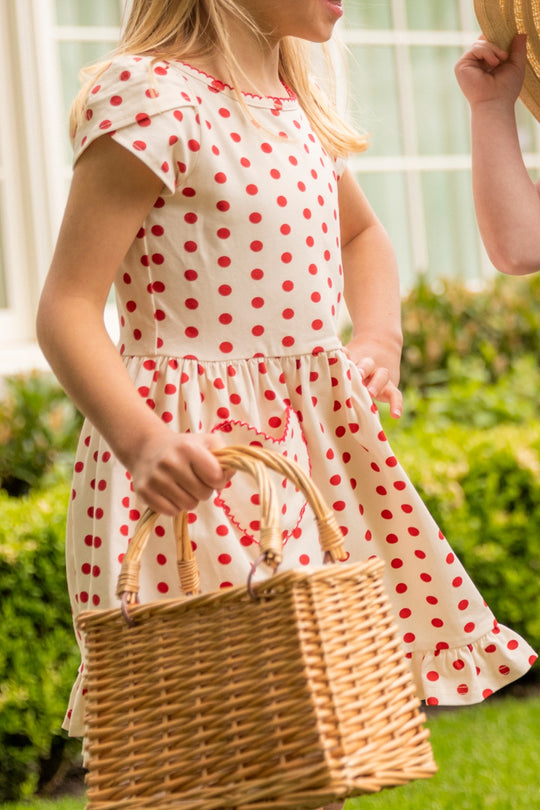 A young girl wears the Ivy City Co Mini Dream Play Dress in Red Polka Dot, holding a woven basket outdoors. Only her upper body shows, with another person's hand partly visible, suggesting a garden scene.