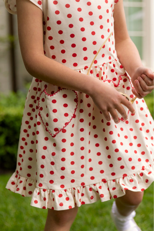A child stands on green grass wearing the Ivy City Co Mini Dream Play Dress in Red Polka Dot, featuring heart-shaped pockets and a ruffled hem. The child’s face is not visible.