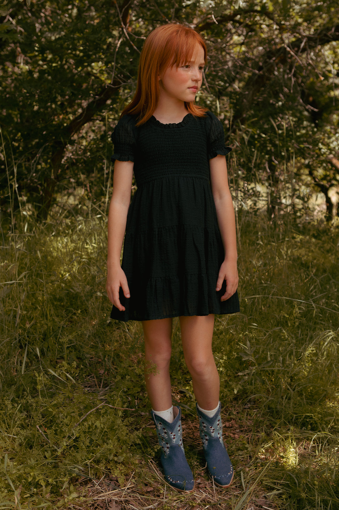 A young red-haired girl stands in tall grass and trees, wearing the Ivy City Co Mini Ellery Dress in Black with blue boots featuring white designs, gazing off to the side in a sunlit forest.
