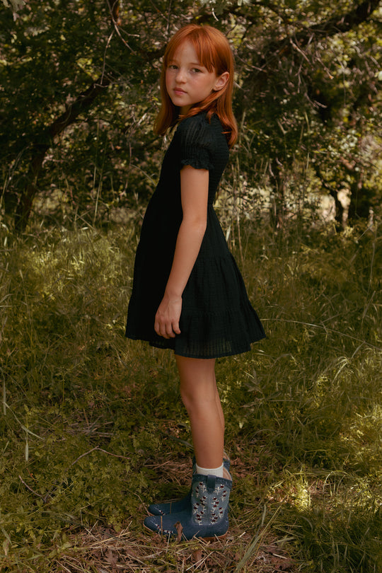A young red-haired girl wears the Ivy City Co Mini Ellery Dress in Black, standing sideways in a grassy, wooded area with blue boots, gazing at the camera as sunlight filters through the trees behind her.