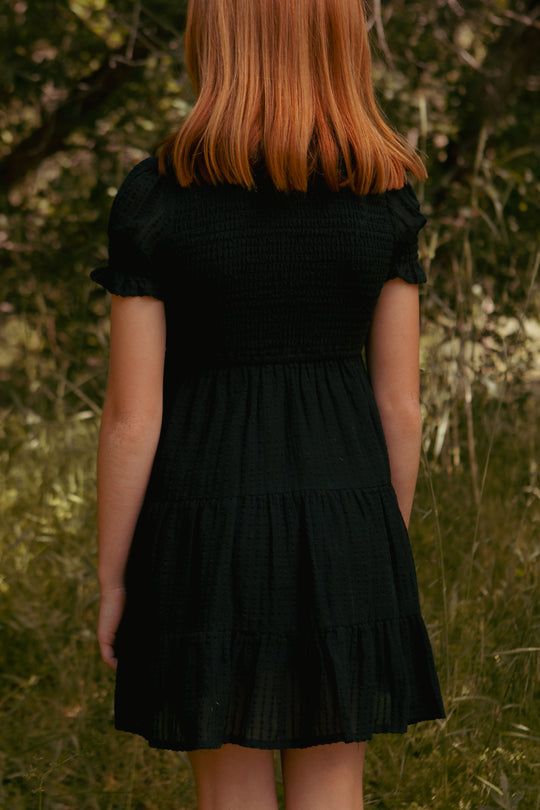 A young girl with straight, shoulder-length red hair stands outdoors, back to the camera, wearing the Ivy City Co Mini Ellery Dress in Black. She is surrounded by green grass and trees.