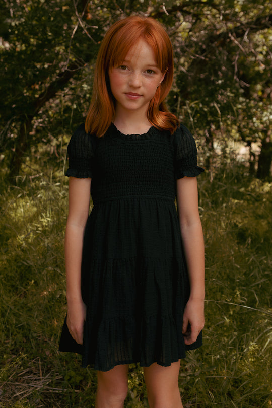 A young girl with red hair stands in a grassy, wooded area wearing the Ivy City Co Mini Ellery Dress in Black. She gazes gently at the camera as sunlight filters through the trees behind her.