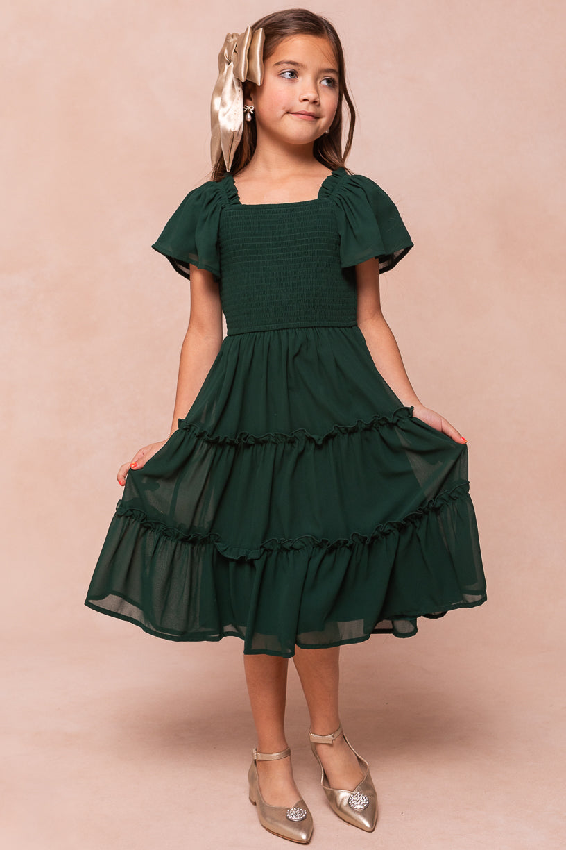 A young girl wears the Ivy City Co Mini Ellie Dress in Emerald Chiffon, featuring a smocked bodice, square neckline, flutter sleeves, and a tiered skirt.