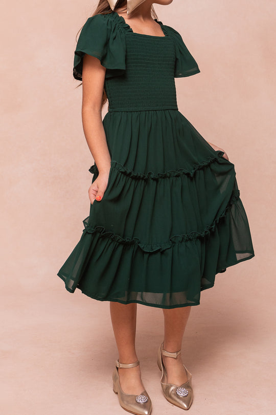 A child wears an emerald chiffon Ivy City Co Mini Ellie Dress, featuring a smocked bodice, flutter sleeves, and a knee-length tiered ruffled skirt.