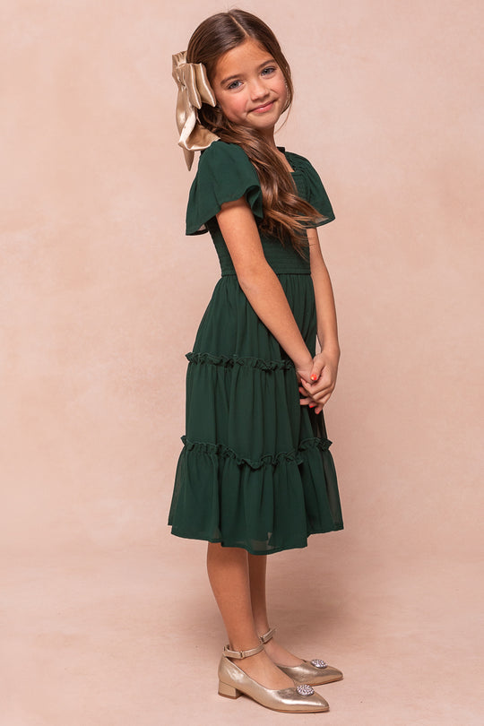 A girl wears an emerald chiffon Mini Ellie Dress from Ivy City Co, featuring a smocked bodice, flutter sleeves, and gold metallic flats.