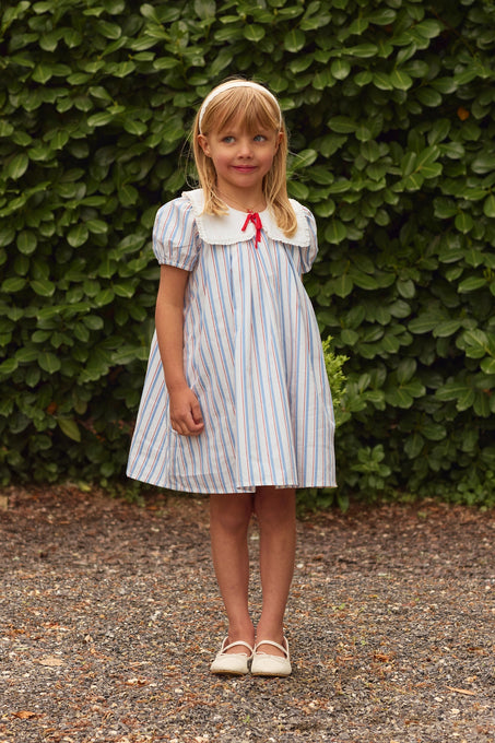 A young girl with blond hair and a headband stands on gravel before green bushes, wearing Ivy City Co’s Mini Emery Dress in Heritage Stripe—a light blue and white girls’ summer dress with a white collar, puff sleeves, red bow, and cream shoes.