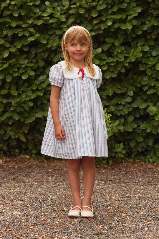 A young girl with blond hair and a headband stands on gravel before green bushes, wearing Ivy City Co’s Mini Emery Dress in Heritage Stripe—a light blue and white girls’ summer dress with a white collar, puff sleeves, red bow, and cream shoes.