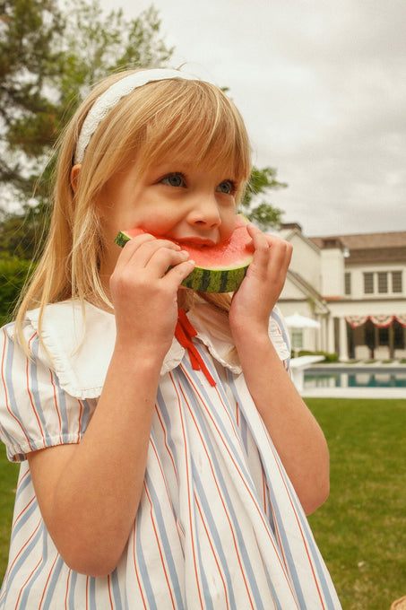 A young girl in the Ivy City Co Mini Emery Dress in Heritage Stripe and a white headband eats watermelon outside, with a large white house and green lawn in the background.