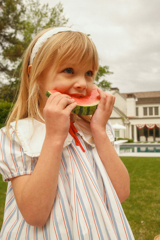 A young girl in the Ivy City Co Mini Emery Dress in Heritage Stripe and a white headband eats watermelon outside, with a large white house and green lawn in the background.