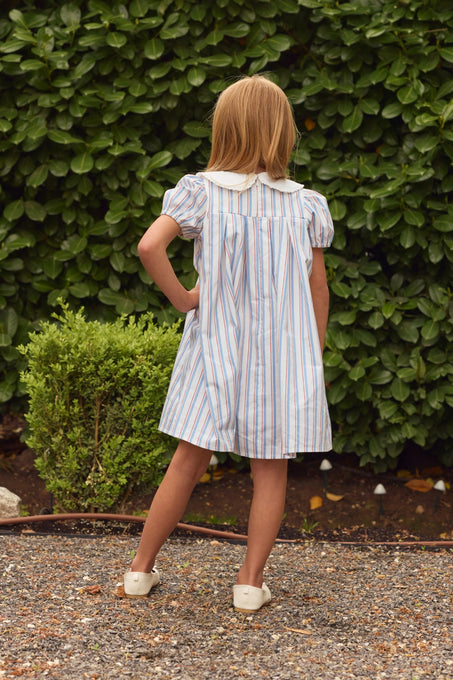A young girl with shoulder-length blonde hair stands outdoors on gravel, facing away, wearing an Ivy City Co Mini Emery Dress in Heritage Stripe and white shoes, with green leafy bushes in the background.