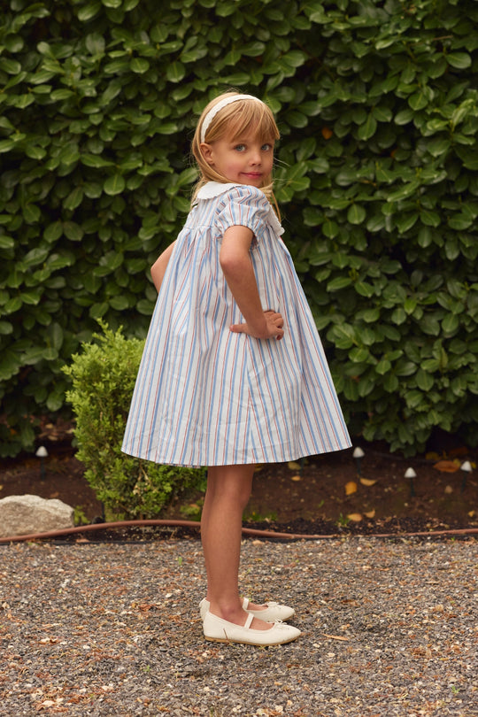 A young girl stands on gravel, hands on hips, wearing Ivy City Co's Mini Emery Dress in Heritage Stripe—white with blue and red stripes—paired with white shoes and a headband. Lush green bushes fill the background.