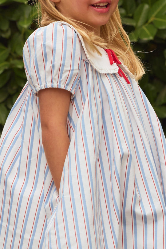 A young girl smiles outside before green foliage, wearing the Ivy City Co Mini Emery Dress in Heritage Stripe— a red, white, and blue girls’ summer dress with short sleeves and red accents.