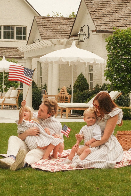 A family of four relaxes on a lawn as parents hold two young children in Ivy City Co's Mini Emery Dress in Heritage Stripe, waving small American flags. They're in front of white houses and outdoor furniture on a sunny day.