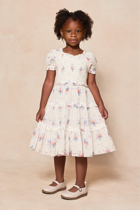 A young girl with curly hair stands against a neutral backdrop, wearing the Ivy City Co Mini Emily Dress in Heritage Soft Ditsy Floral with short puff sleeves and white Mary Jane shoes—a sweet look for mom and me matching moments.