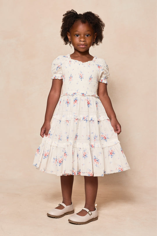 A young girl with curly hair stands against a neutral backdrop, wearing the Ivy City Co Mini Emily Dress in Heritage Soft Ditsy Floral with short puff sleeves and white Mary Jane shoes—a sweet look for mom and me matching moments.