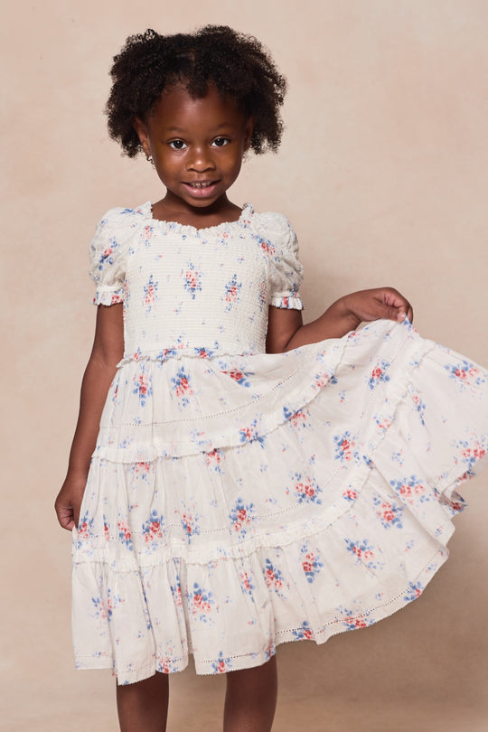 A young girl smiles as she holds out the skirt of her Mini Emily Dress in Heritage Soft Ditsy Floral by Ivy City Co, featuring puffed sleeves—perfect for mom and me matching outfits against a beige backdrop.